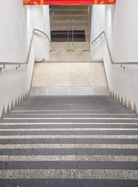 A dimly lit stairwell leading down to a basement or lower level, featuring dark, textured concrete steps with a metal handrail on the left side. The surrounding walls are painted in a muted, uneven gr