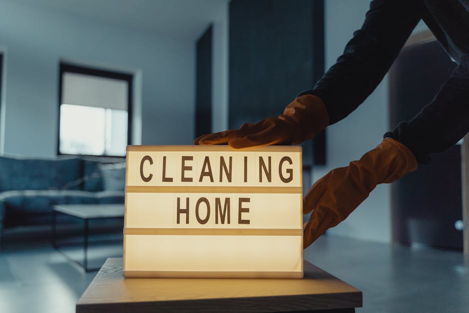 A person wearing brown cleaning gloves is adjusting a lightbox sign that displays the words 'CLEANING HOME' in black letters. The sign is illuminated from within, placed on a wooden surface, and posit
