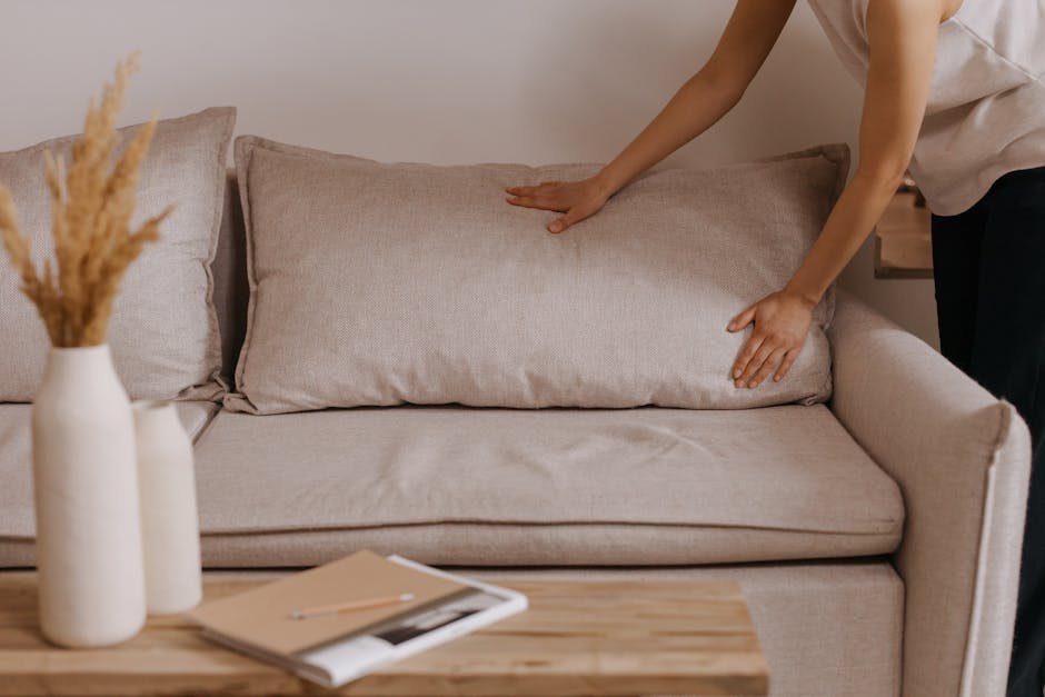 A person wearing a light-colored sleeveless top is smoothing the beige fabric cushion on a light-colored fabric sofa in a living room. The sofa has a smooth, clean surface with no visible dust or stai