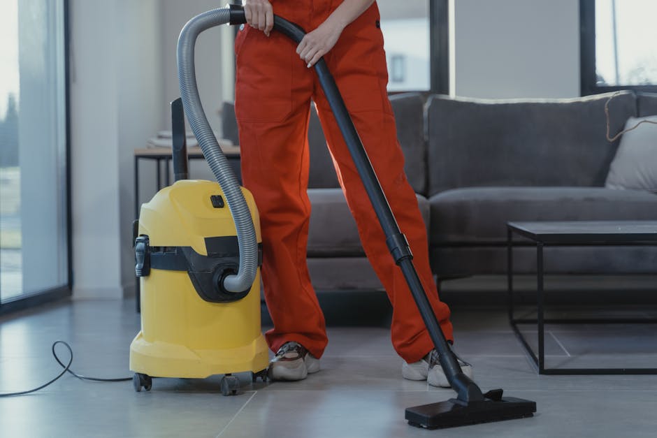 A person wearing orange work trousers and grey shoes is operating a yellow vacuum cleaner with a grey hose in a modern living room. The vacuum cleaner is positioned on a tiled floor, and the person is